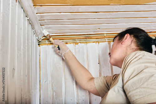 Coating edge, female painter applying white layer along meeting line of vertical and horizontal wood paneling to achieve consistent interior finish.