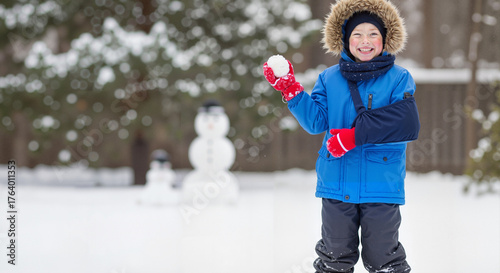 Happy boy with a broken arm in a sling playing with a snowball in winter. Cheerful child enjoying the snow despite an injury. Childhood resilience and recovery concept with copy space