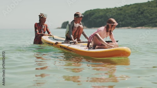 Mother and children playing on SUP board in calm sea on sunny summer day. Family vacation and outdoor fun together by ocean on paddleboard.