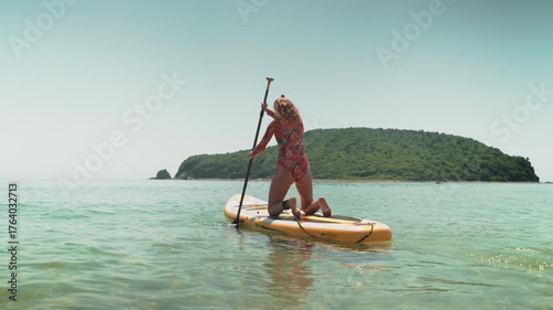 Sporty woman enjoying summer day paddling on yellow board in turquoise sea. Concept of travel, leisure, and joyful freedom on water. Summer holidays, freedom, and privacy.