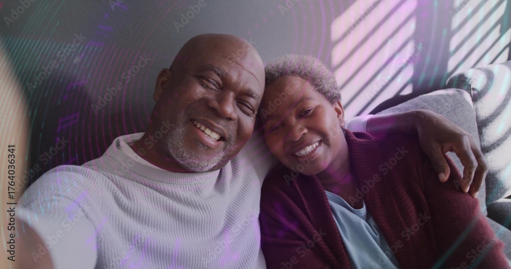 Fototapeta premium Smiling senior couple leaning together taking selfie on light sofa, with window blinds shadows