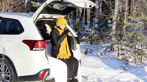 Happy girl drinking hot tea while sitting in the trunk of a car. Winter travel by car.