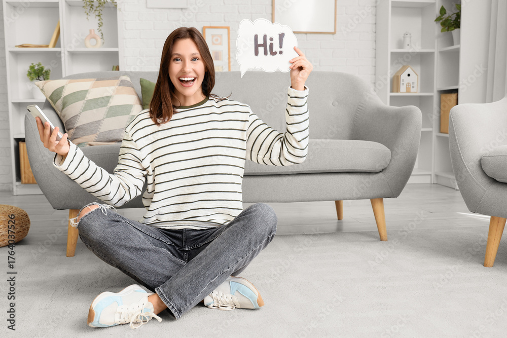 © Pixel-Shot - Young woman holding speech bubble with word HI and mobile phone at home