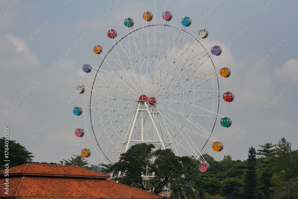 Fototapeta premium A large Ferris wheel with colorful cabins set against a cloudy blue sky and trees.