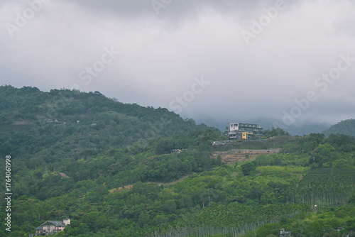 Green mountain hillside with white buildings on cleared red soil platform viewed from Pobuwo embankment, Datong Township mountains across Lanyang River under overcast sky, Yilan.