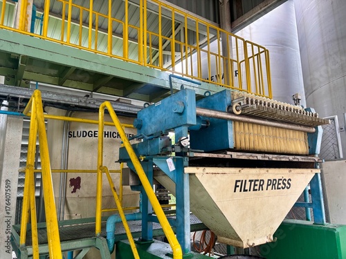 A large Filter Press machine in a wastewater treatment facility, clearly showing the dewatering mechanism and collection hopper, with yellow railings and a Sludge Thickener sign in the background.