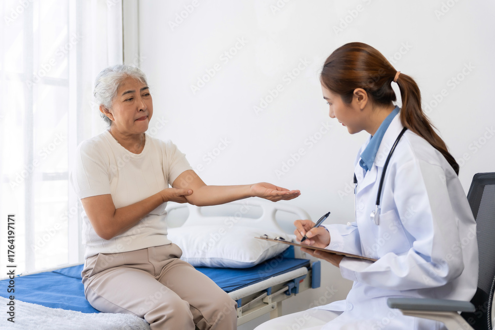Fototapeta premium Senior woman experiencing elbow pain, consulting with a female doctor who is writing notes on a clipboard in a medical room