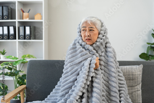 Asian senior woman feeling cold, shivering, and suffering from flu symptoms or fever while wrapped in a warm blanket at home