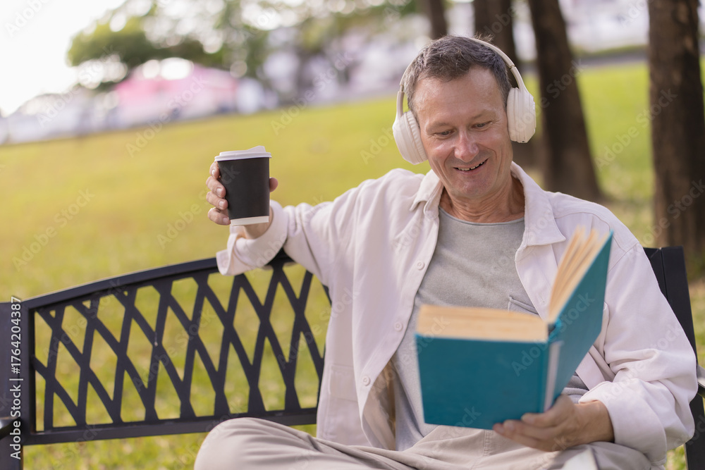 Fototapeta premium Senior man on a park bench enjoying a quiet afternoon: reading a book, sipping coffee and listening to music with headphones, relaxed and content in nature
