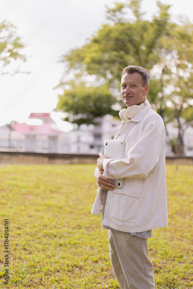 Obraz premium Senior man standing outdoors in a park, smiling and holding a modern digital tablet while wearing wireless headphones around his neck, reflecting a tech-savvy lifestyle