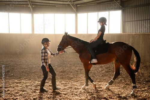 Obraz na plátně Riding instructor teaches teenage girl wearing riding helmet how to ride horse in indoor arena