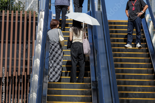 Two friends ride escalator up towards open air plaza with blue sky backdrop. Both have shoulder bags, and one shelters under sun umbrella. Man riding downwards stands casually on adjacent escalator.