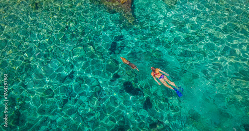 AERIAL TOP DOWN: Woman with snorkelling fins swims with her dog in shimmering sea. Incredible clarity of Adriatic Sea allows sunlight to illuminate textured seabed. Unspoiled nature on island of Hvar
