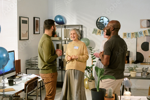 Senior Caucasian woman smiling and talking with middle aged Caucasian man and Black man during retirement celebration in office setting, people standing and interacting near decorations