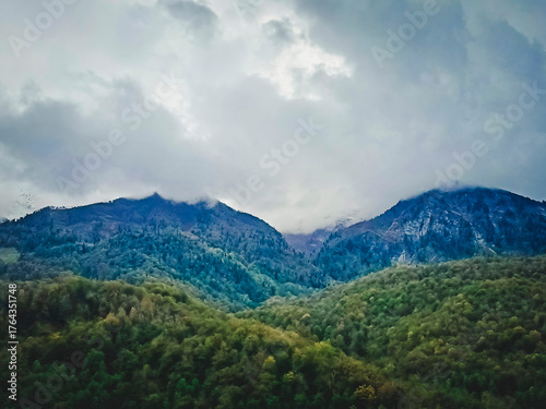 Aerial view of the majestic Caucasus Mountains in a haze, a green valley surrounded by forested mountains under a cloudy sky.