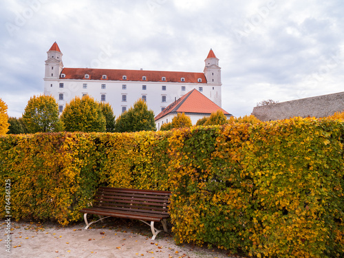 Photography Bratislava Castle with autumn linden trees and trimmed hedges in a quiet baroque garden, with an empty bench creating a peaceful atmosphere