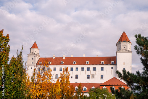 Photography Bratislava Castle in Slovakia with red roofs and white walls, surrounded by autumn trees and flowers under a cloudy sky
