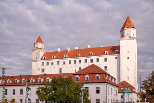 Photography Bratislava Castle in Slovakia with red roofs and white walls, surrounded by autumn trees and flowers under a cloudy sky