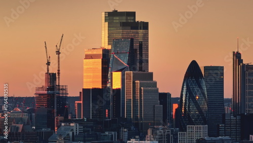 Bright orange sunset light over modern glass skyscrapers in London's financial district, iconic buildings like the Gherkin and active construction cranes. Business center skyline. Drone flight