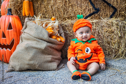 A little boy wearing a pumpkin costume and hat sits on a woven cloth to celebrate Halloween with a background of pumpkins, ghosts and skeletons.
