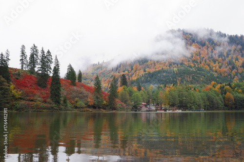 A view across a calm lake of a lakeside building and hills covered in colorful red and orange autumn foliage under a cloudy sky