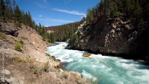 Dynamic action shot of a team whitewater rafting through turbulent rapids in a rocky river gorge. Paddlers wearing bright helmets and lifejackets power an inflatable raft with paddles, splashing water
