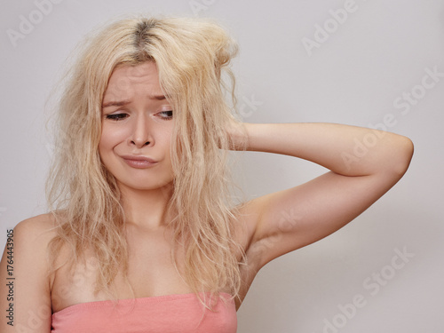 Young blonde woman with messy hairstyle and confused expression holding her head, representing stress, frustration and everyday emotional challenges in beauty lifestyle. Bleached hair, emotional face