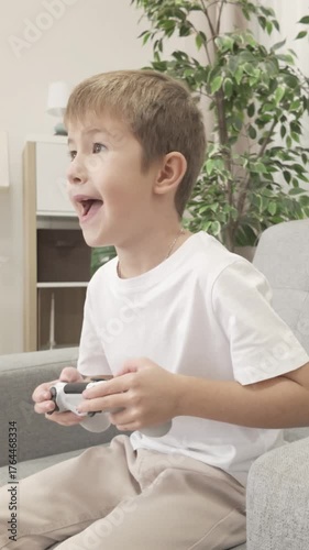 Seven year old boy sitting in armchair and playing video game with joystick, showing joy and excitement after winning