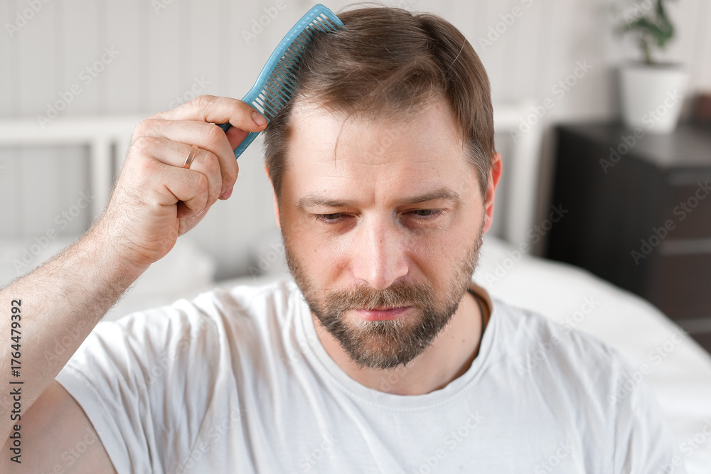 Obraz premium Handsome man combing brushing hair sitting at home light white bedroom. Guy styling grooming his hair with a comb. Self-care. Softly lit indoor space. Portrait. Male haircare morning routine concept
