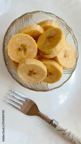 Closeup of fork and organic ripe banana fruit cut piece slice serve on glass bowl isolated on white plate background. Healthy eating, weight loss diet, wellness lifestyle and snack craving concept.