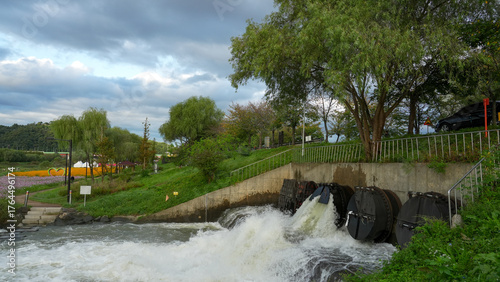 water flow from floodgate with trees and flower field under cloudy sky