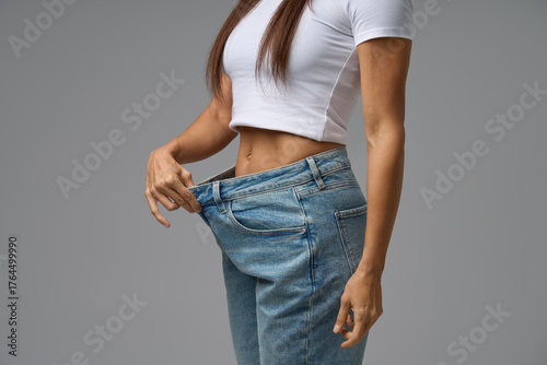 Fit woman in white crop top showing significant weight loss by holding loose blue jeans on gray background
