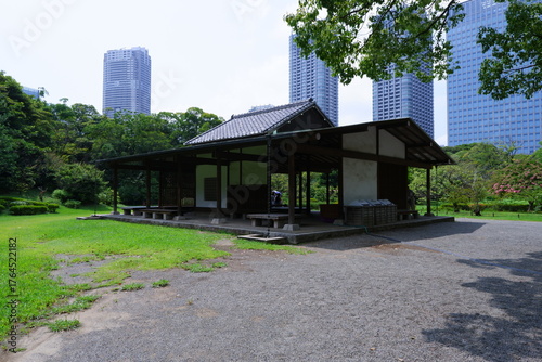 A traditional wooden teahouse or rest shelter in a Japanese public garden, with modern high-rise buildings in the background