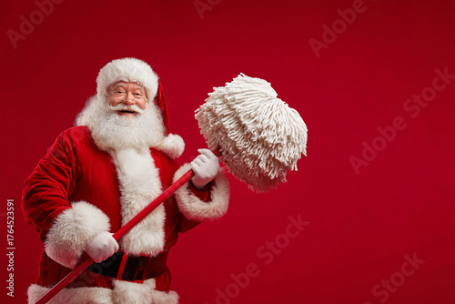 A cheerful Santa Claus in a red suit with a huge fluffy beard holds a large mop in his hands.