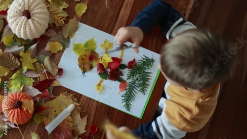 Sweet child, boy, applying leaves using glue while doing arts and crafts at home, autumntime
