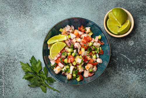 Fresh and vibrant peruvian seafood ceviche with shrimps, octopus, tomatoes, cucumber and parsley, served with lime in a bowl on a stone background from above, dish of Peru