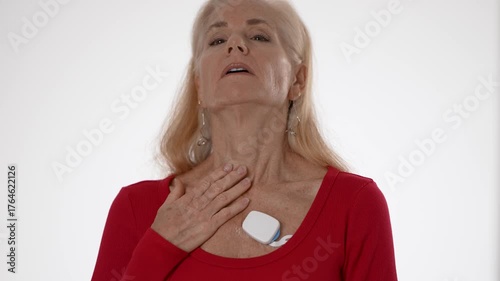 A happy mature woman feeling her heart using a heart sensor to keep track of her atrial fibrillation. She stands isolated on a white background, showcasing health monitoring.