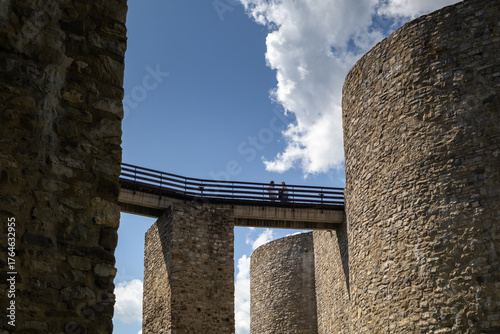 historical stone castle towers connected by a wooden bridge under a bright blue sky with white clouds, showcasing ancient medieval architecture and two people walking across
