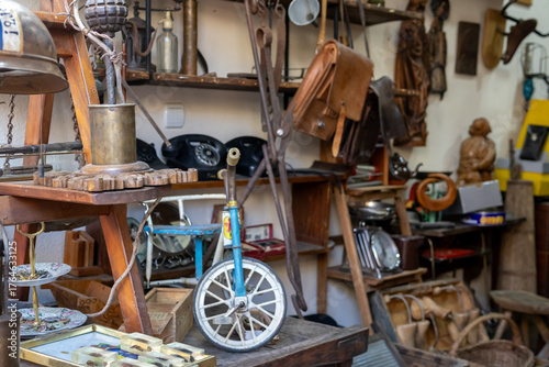 eclectic vintage items crowd a dusty antique shop or flea market stall, featuring a small blue unicycle, leather bags, and various retro curiosities
