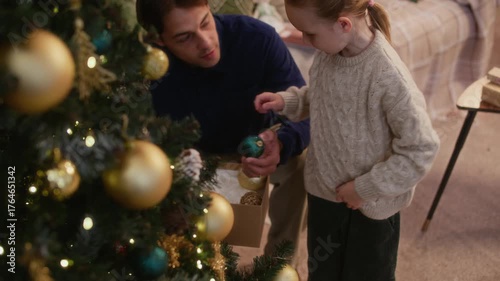 Loving Caucasian father and his little daughter hanging ornaments on Christmas tree and sharing warm conversation while enjoying joyful holiday moments together in warm, cozy home setting