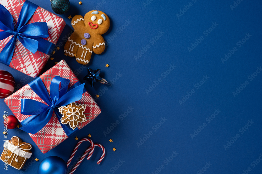 Fototapeta premium A festive arrangement of wrapped gifts with blue ribbons, gingerbread man, cookies, and candy canes on a blue backdrop, suggesting Christmas and New Year celebration and warm holiday greetings