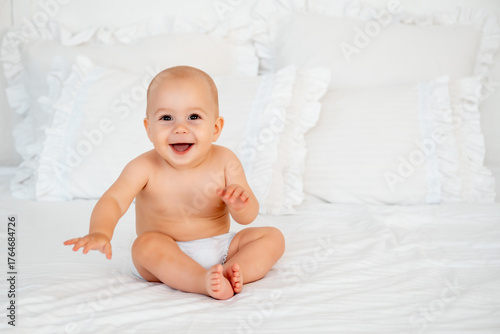 a baby in a diaper on a white bed, space for text, a small, smiling child sitting on a bed at home