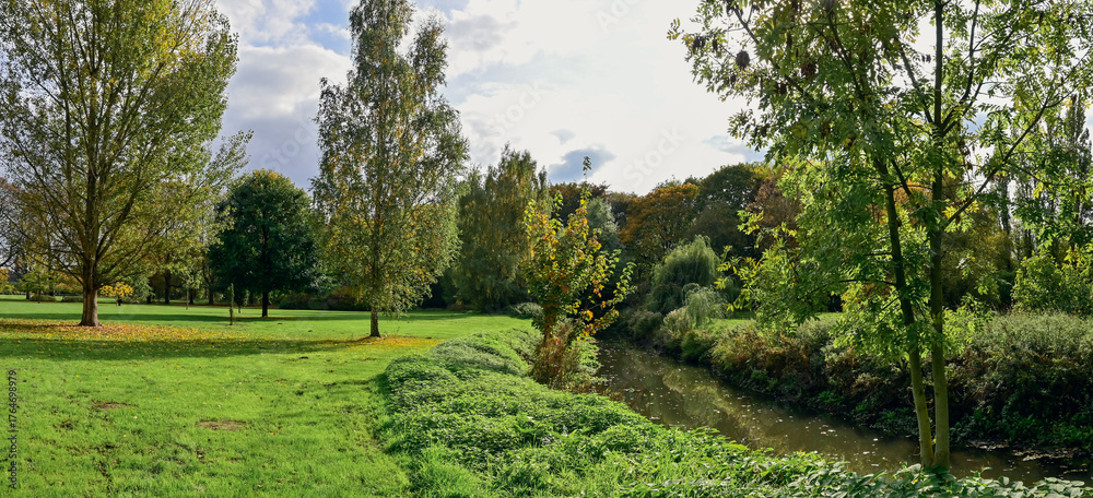 Naklejka premium Panoramic landscape photograph of the Rivierenhof in Deurne (Belgium) with grass, green trees, river and sky with clouds.