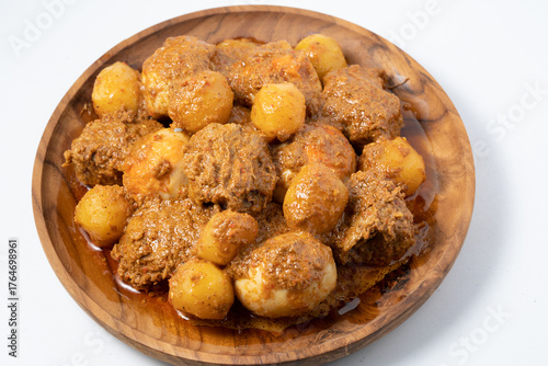 A top-down view of a wooden plate holding beef, hard-boiled eggs, and small potatoes cooked in a thick, brown Indonesian rendang sauce. Home cooking style