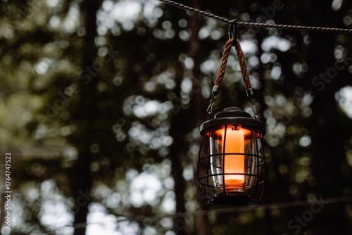 A glowing vintage lantern hanging by a rope in the quiet forest twilight. Captured with a shallow depth of field, the warm light contrasts beautifully against the dark, moody background of trees