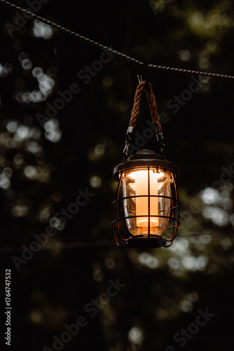 A glowing vintage lantern hanging by a rope in the quiet forest twilight. Captured with a shallow depth of field, the warm light contrasts beautifully against the dark, moody background of trees