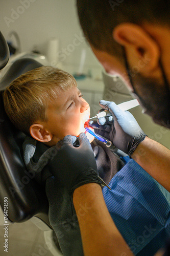 Little boy at dentist appointment sitting in chair while dentist uses dental drill and saliva ejector during treatment. Pediatric dentistry and oral care concept.