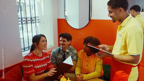 Friendly waiter using a digital tablet to take an order from a diverse group of smiling friends sitting together at a table, discussing the menu and enjoying their time in a vibrant cafe