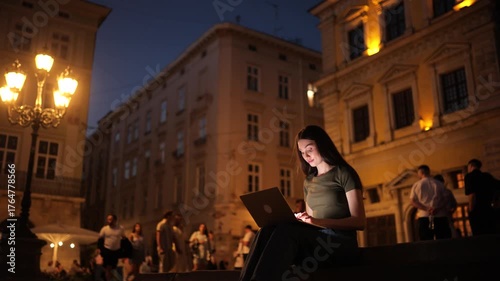 Young woman working on a laptop outdoors at night, illuminated by screen light, sitting on city steps surrounded by warm street lamps and evening crowds
