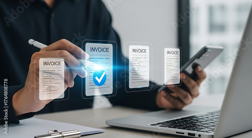 Businessman uses a stylus to approve digital invoices on a virtual interface displayed above a laptop, with a smartphone in his other hand.